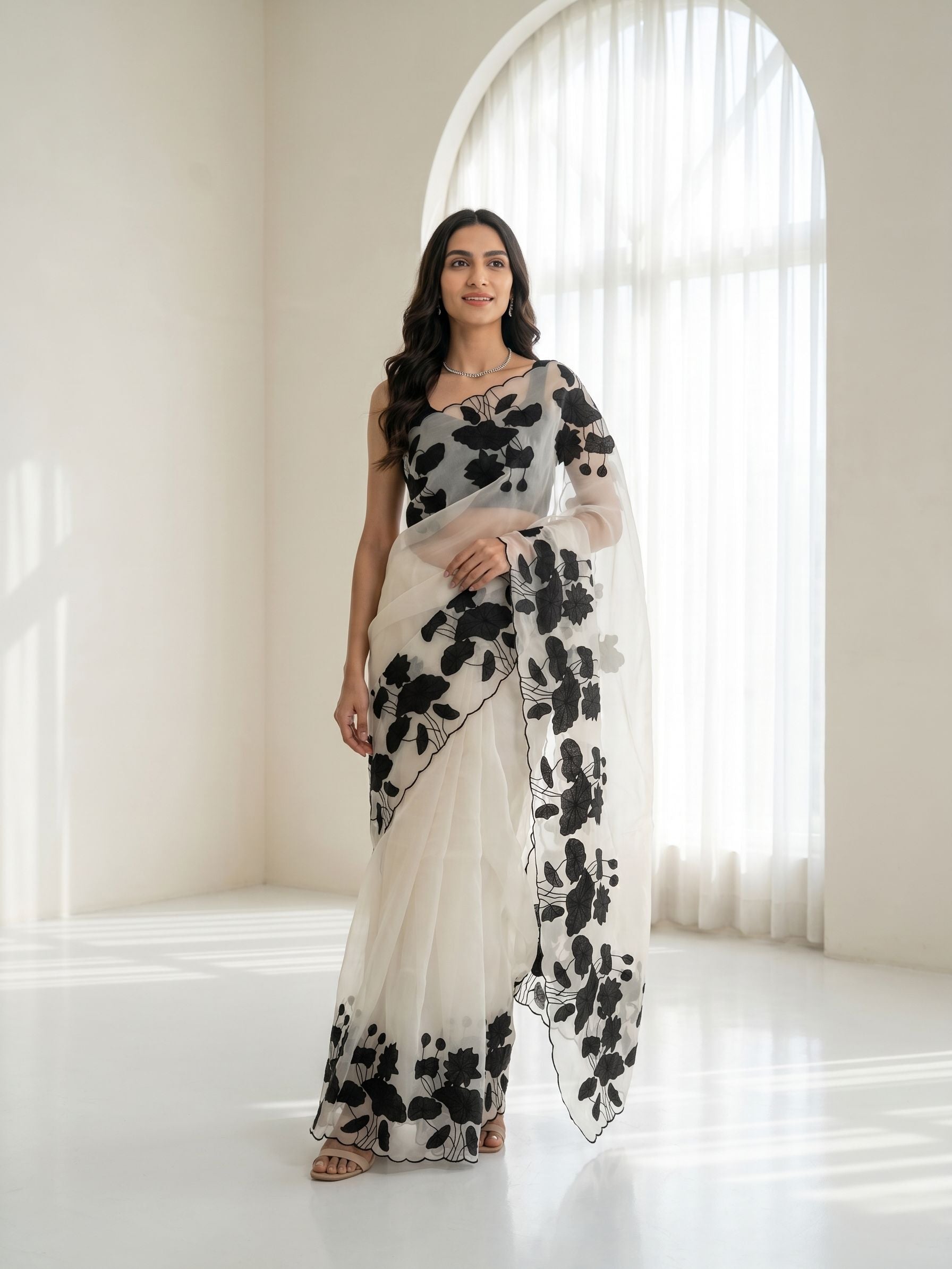 Woman wearing a white saree with black floral patterns in a bright room.
