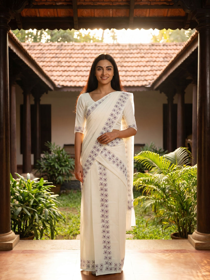 Woman in a white saree with black patterns standing in a traditional setting with greenery.