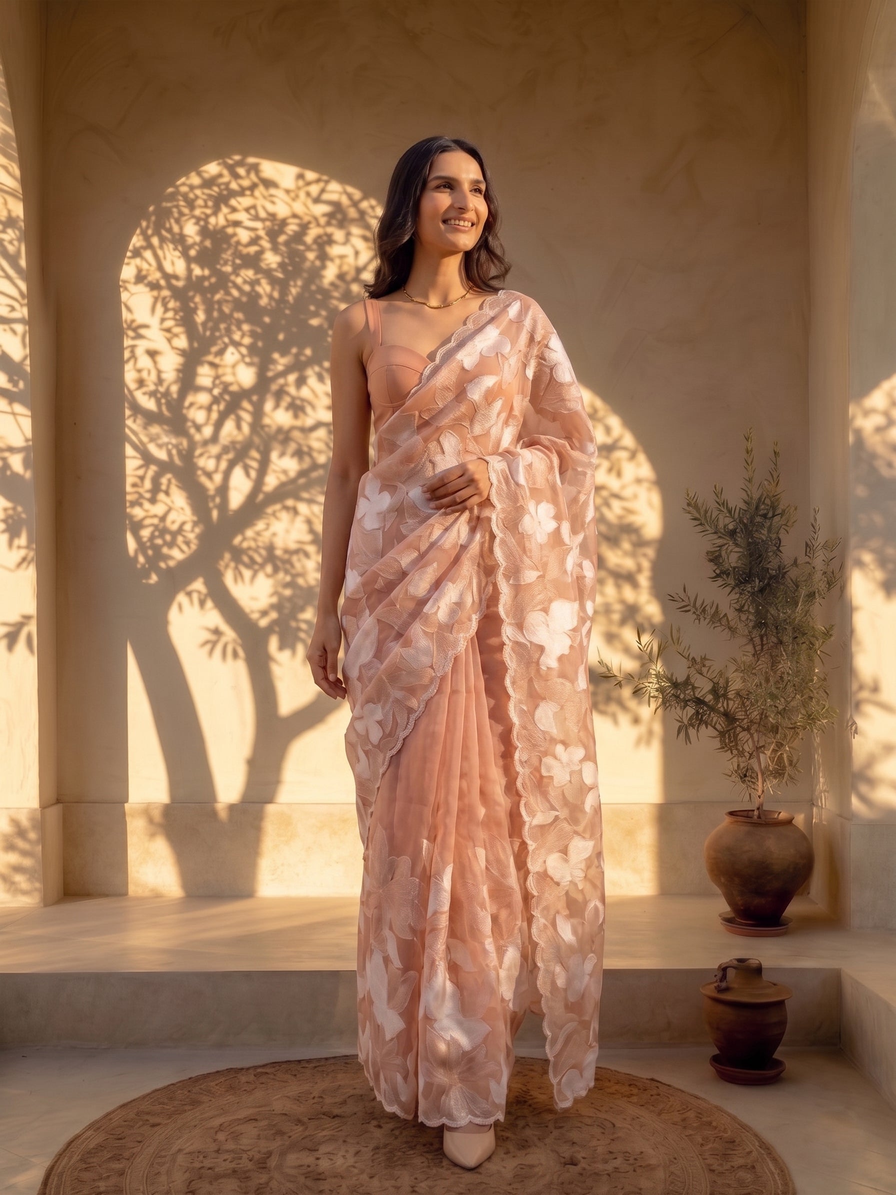 Woman in a floral saree standing in a sunlit room with shadows on the wall.