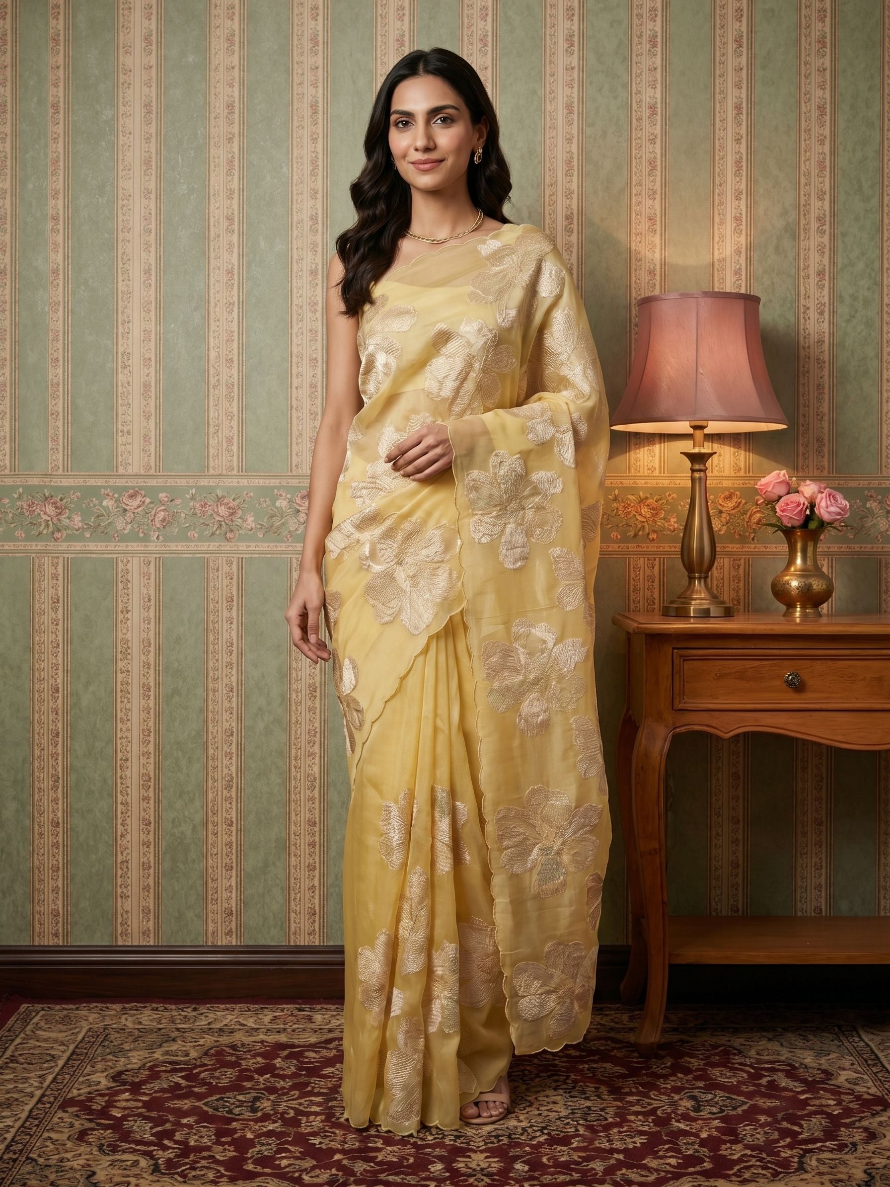 Woman in a yellow saree standing in a room with a lamp and vase on a table.