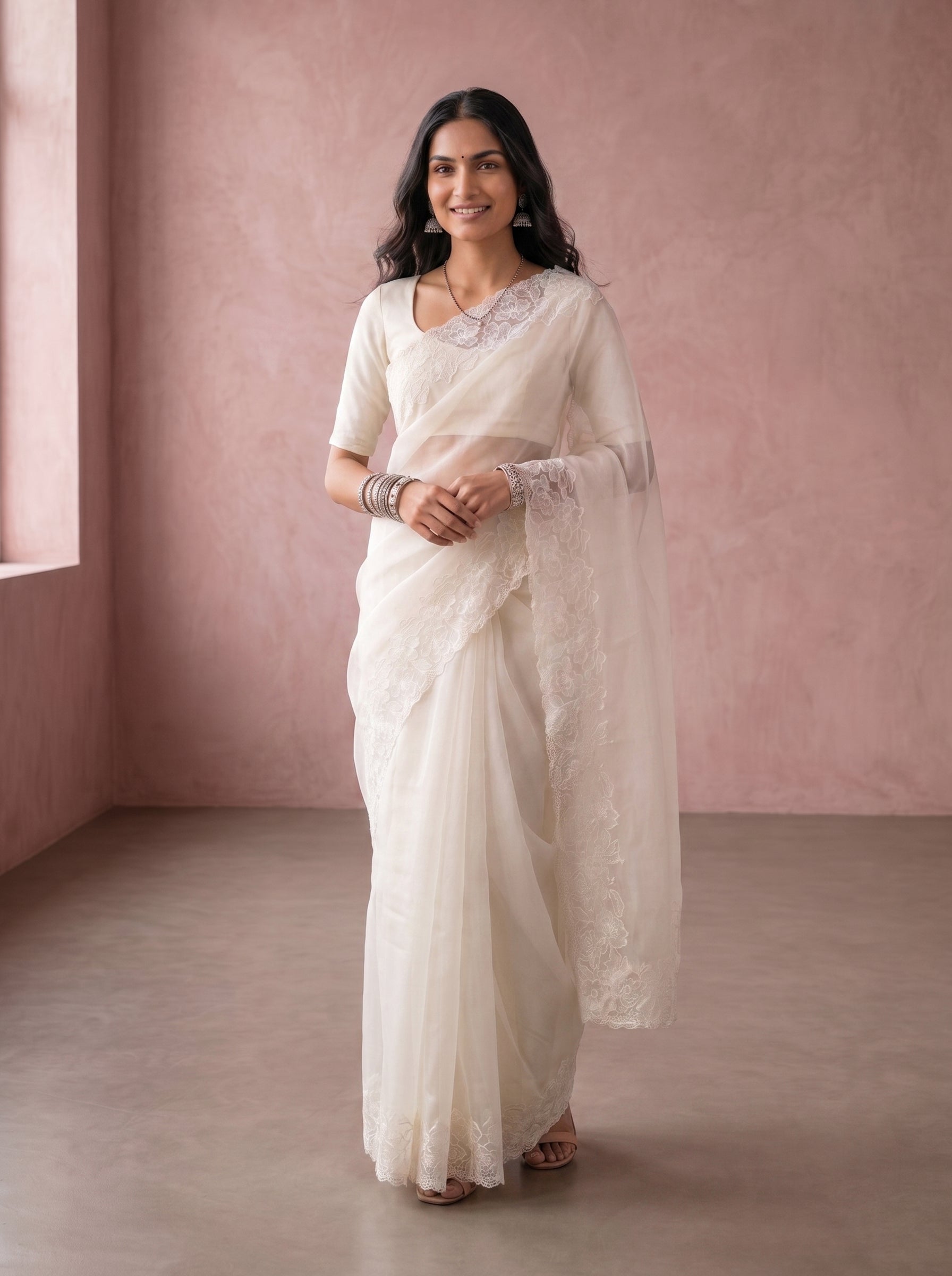 Woman in a white saree standing in a room with pink walls.