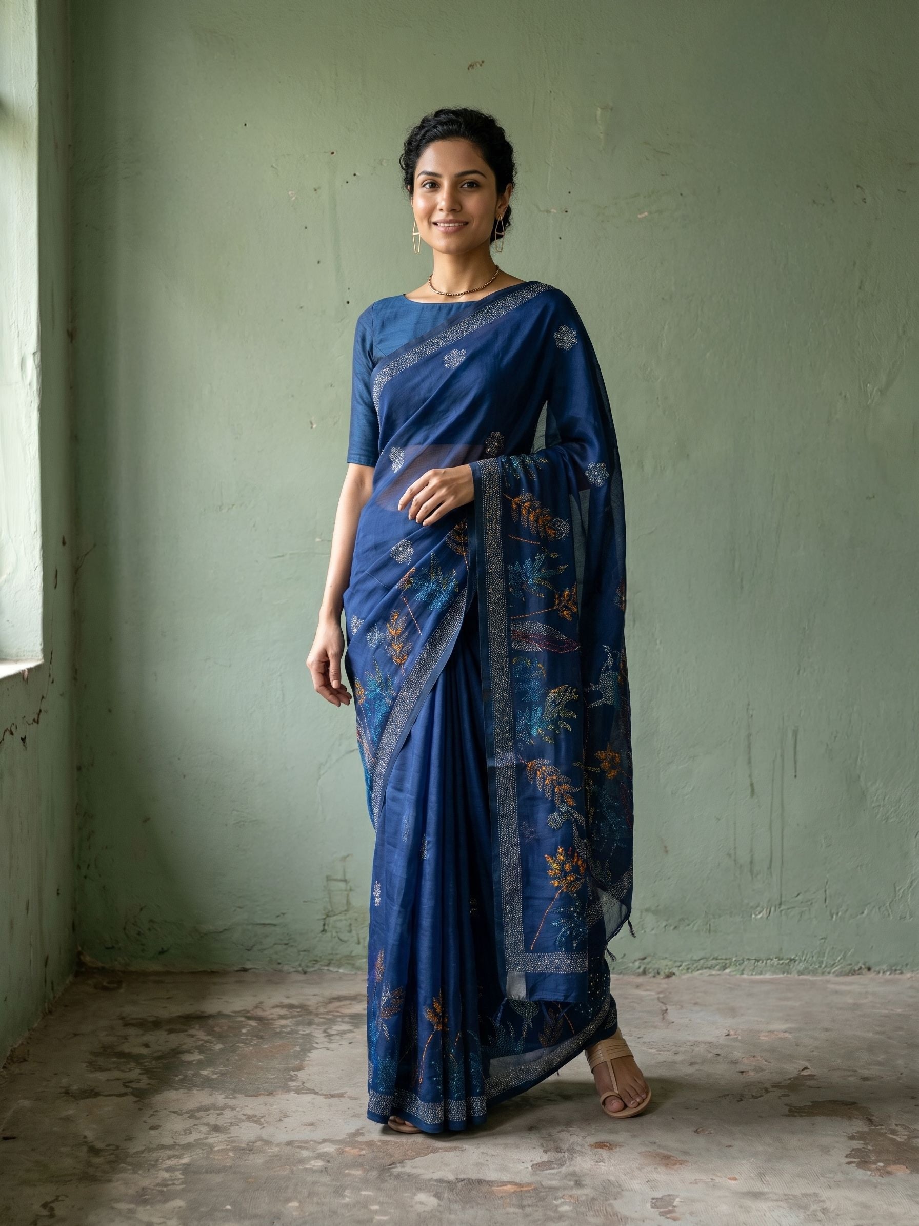 Woman wearing a blue saree with a subtle pattern against a plain wall.
