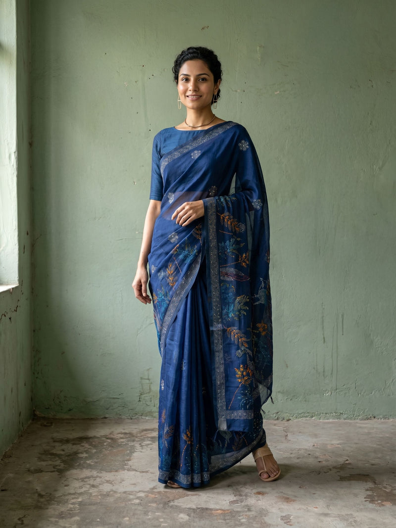Woman wearing a blue saree with a subtle pattern against a plain wall.