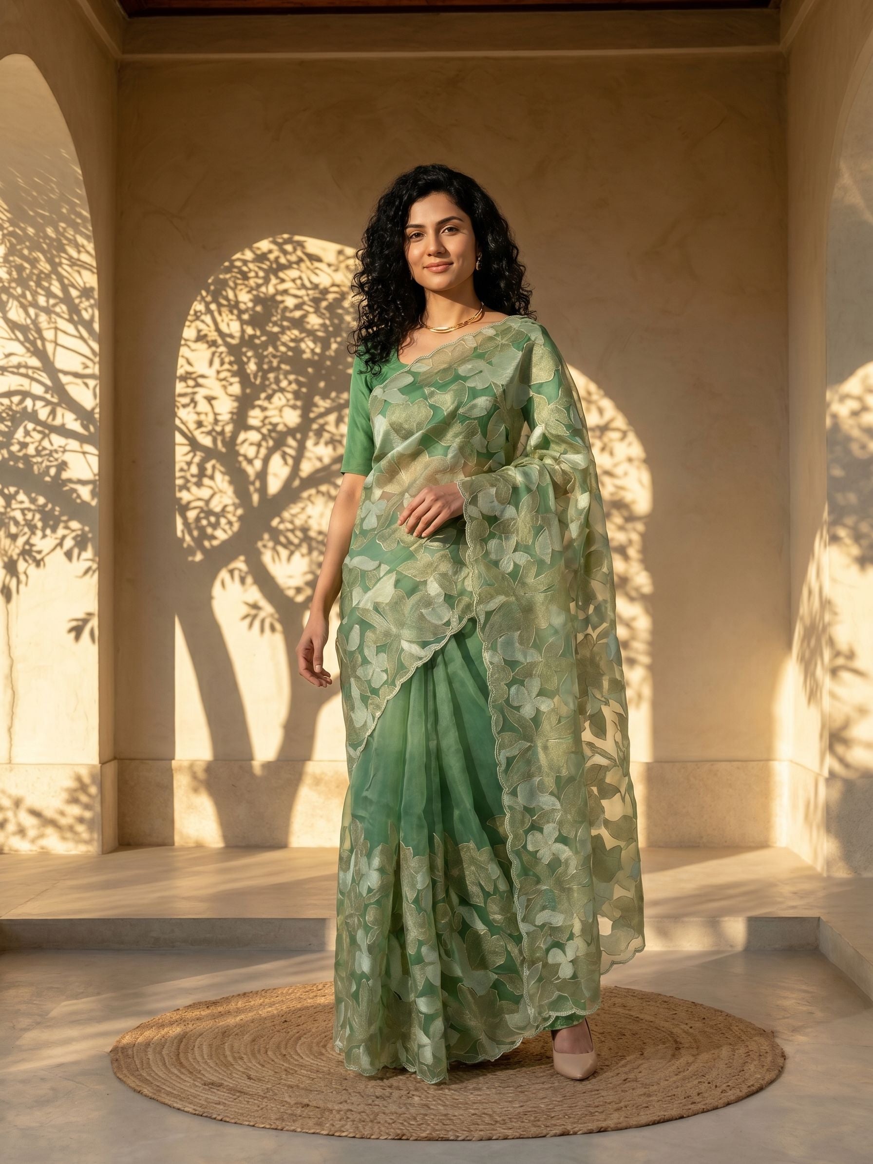 Woman in a green floral saree standing in a sunlit room with shadows on the wall.