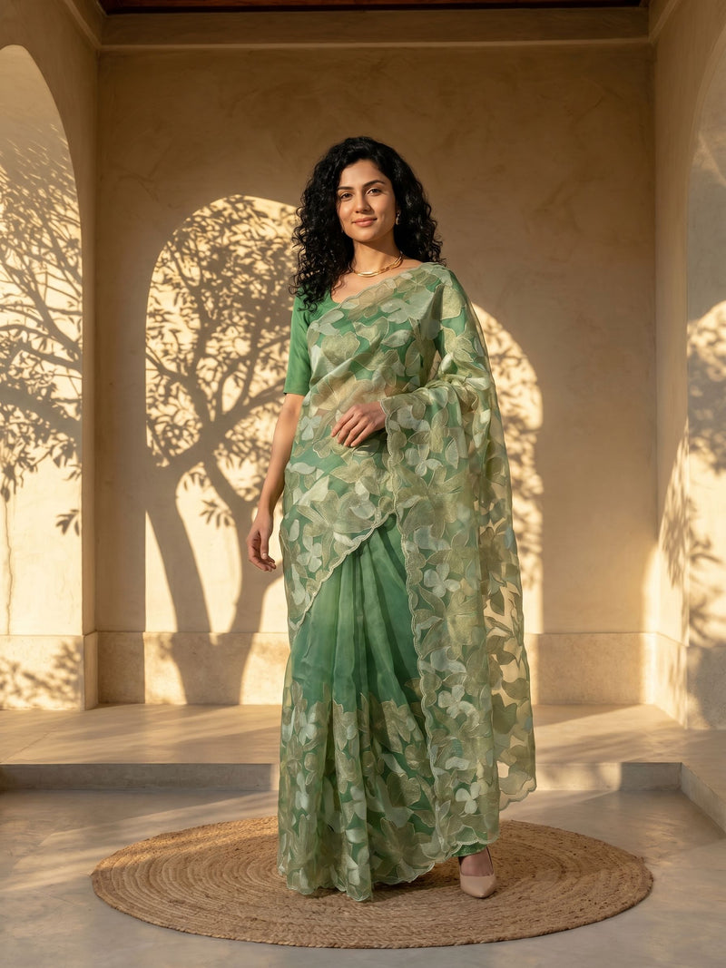Woman in a green floral saree standing in a sunlit room with shadows on the wall.
