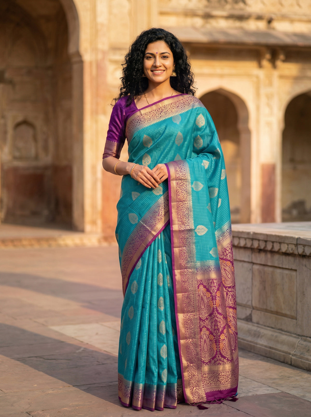 Woman in a turquoise saree with purple border standing in front of an architectural background