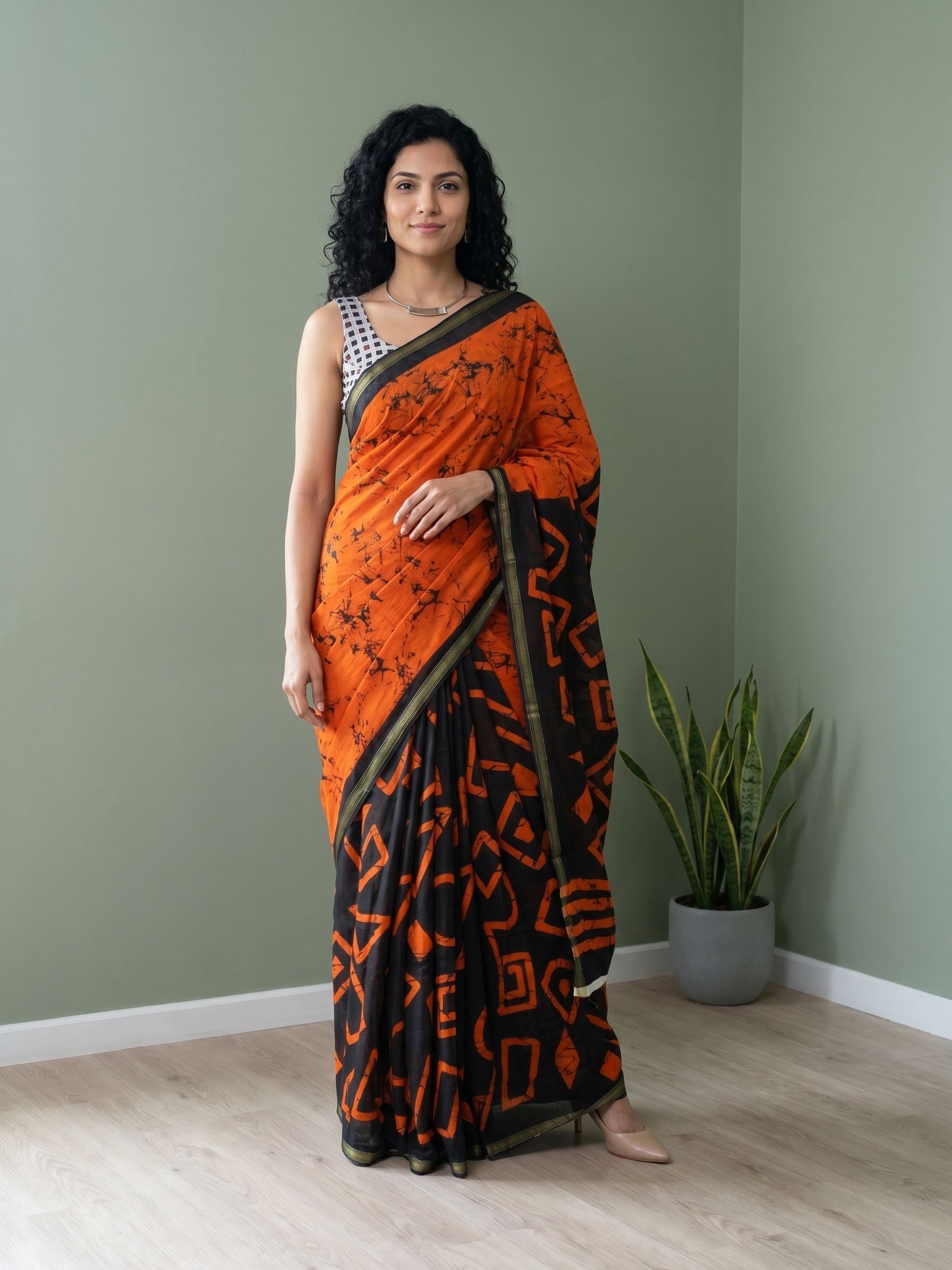 Woman wearing an orange and black saree standing in a room with a plant.