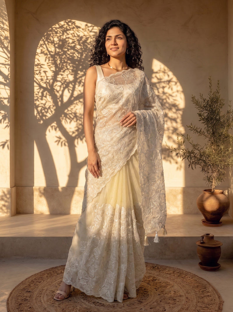 Woman in a white saree standing in a sunlit room with shadows on the wall.