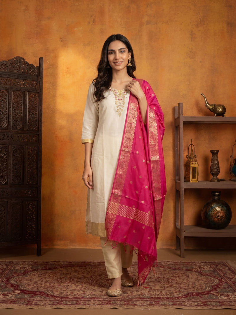 A woman models a traditional Indian outfit consisting of a cream kurta with gold embroidery, beige trousers, and a vibrant pink dupatta with gold zari borders, set against a rustic orange wall with wooden shelves.