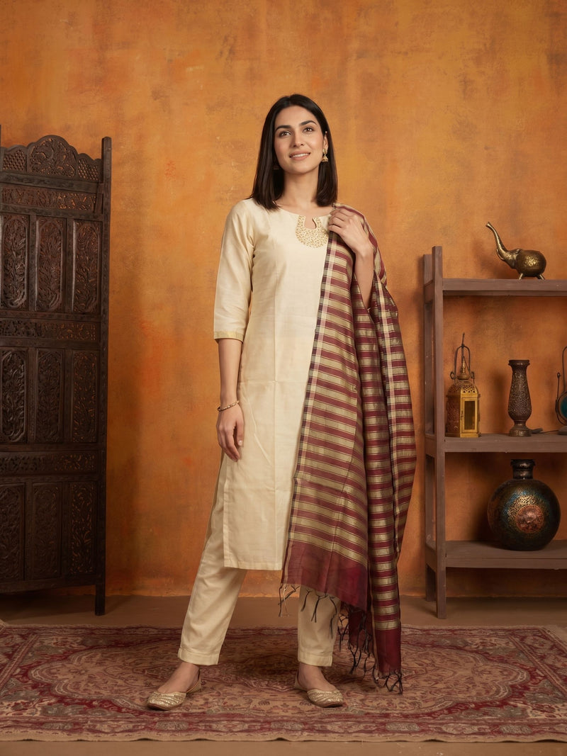 A woman modeling a cream-colored kurta and trouser set with a maroon and beige checkered dupatta, standing in a warm, rustic room with an orange textured wall, wooden furniture, and a patterned rug.