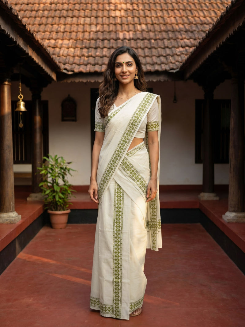 Woman in a white saree with green patterns standing in a traditional setting.