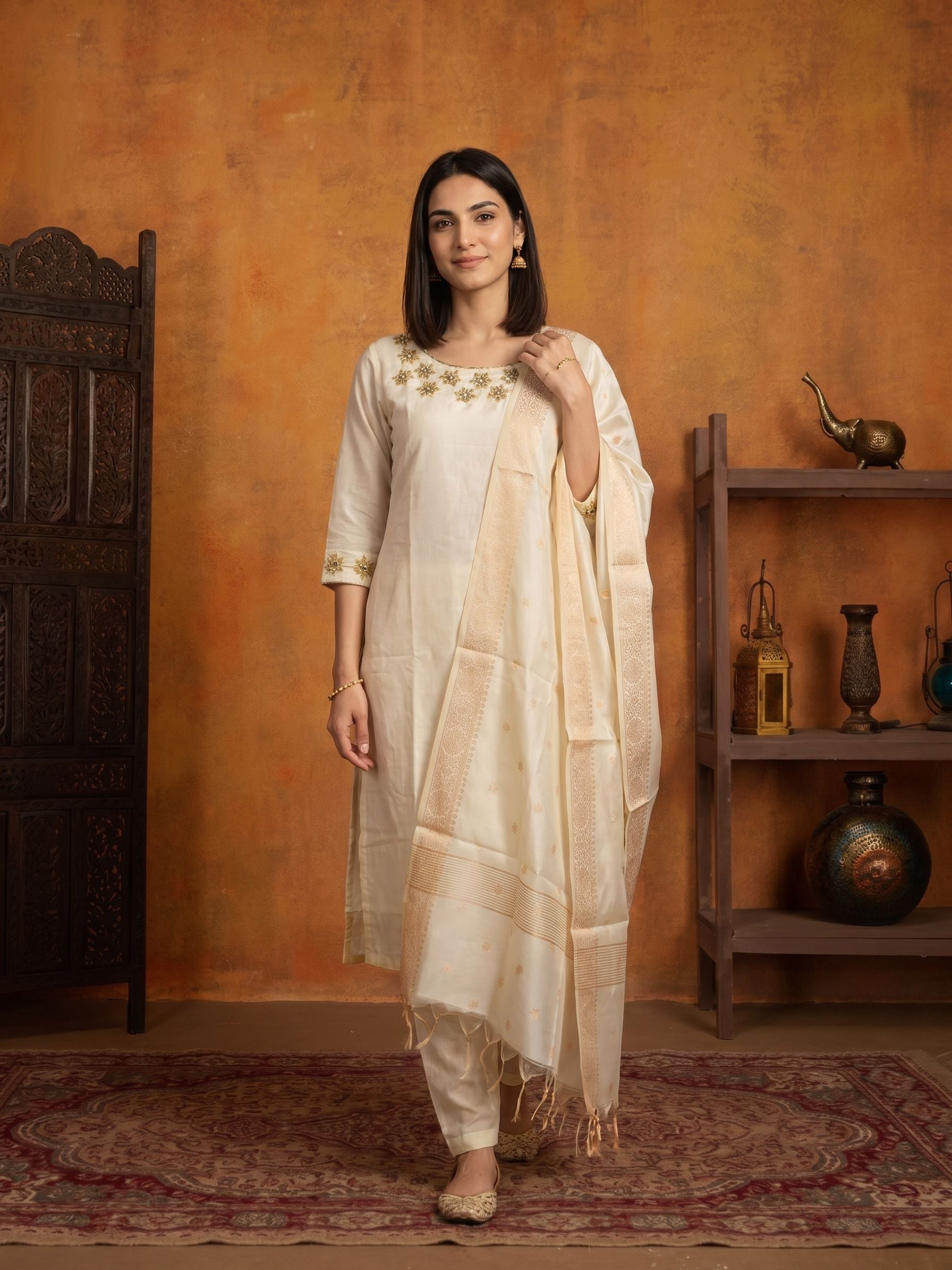 A woman models a traditional Indian outfit consisting of a cream-colored kurta with gold embroidery, matching trousers, and a sheer cream dupatta, set against a rustic orange wall with a wooden shelf and carved door.