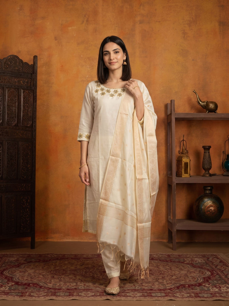A woman models a traditional Indian outfit consisting of a cream-colored kurta with gold embroidery, matching trousers, and a sheer cream dupatta, set against a rustic orange wall with a wooden shelf and carved door.