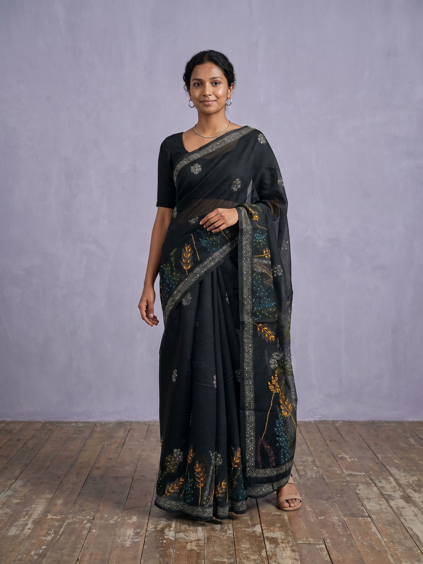 Woman wearing a black saree with floral patterns on a wooden floor and gray wall background