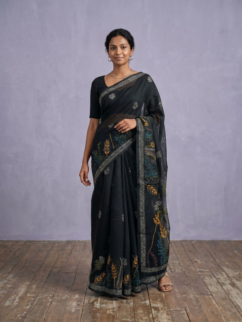 Woman wearing a black saree with floral patterns on a wooden floor and gray wall background
