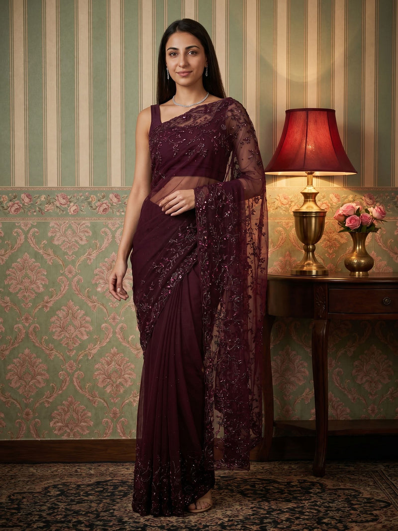 Woman in a dark purple saree standing in a room with decorative wallpaper and a lamp.