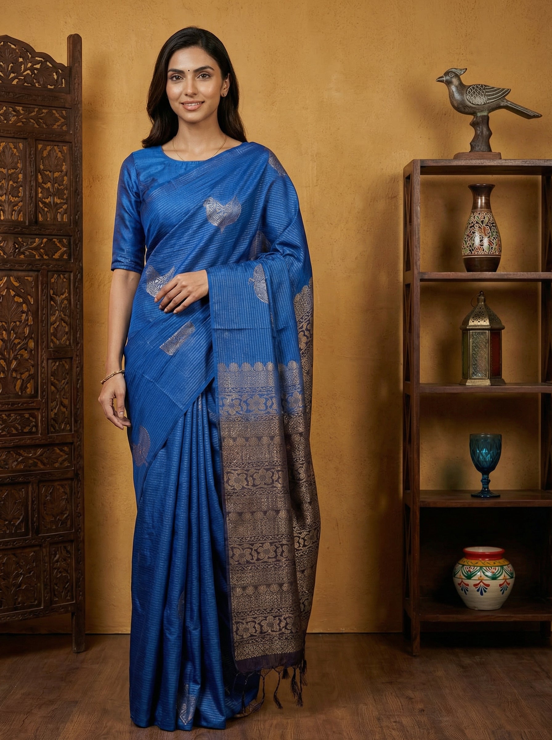 Woman in a blue saree standing in a room with wooden furniture and decorative items.