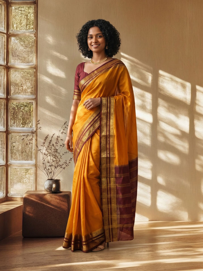 Woman in a yellow saree with a maroon blouse standing in a room with sunlight streaming through a window.
