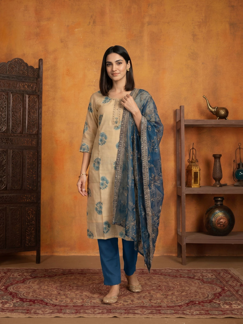A woman models a traditional Indian outfit consisting of a beige kurta with blue floral prints, blue pants, and a blue patterned dupatta, set against a warm orange wall with a wooden screen and shelf.