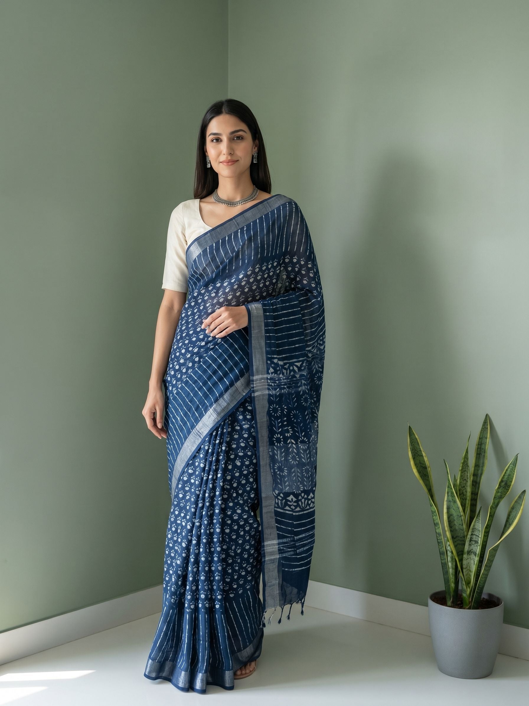 Woman wearing a blue saree standing against a green wall with a plant on the right.