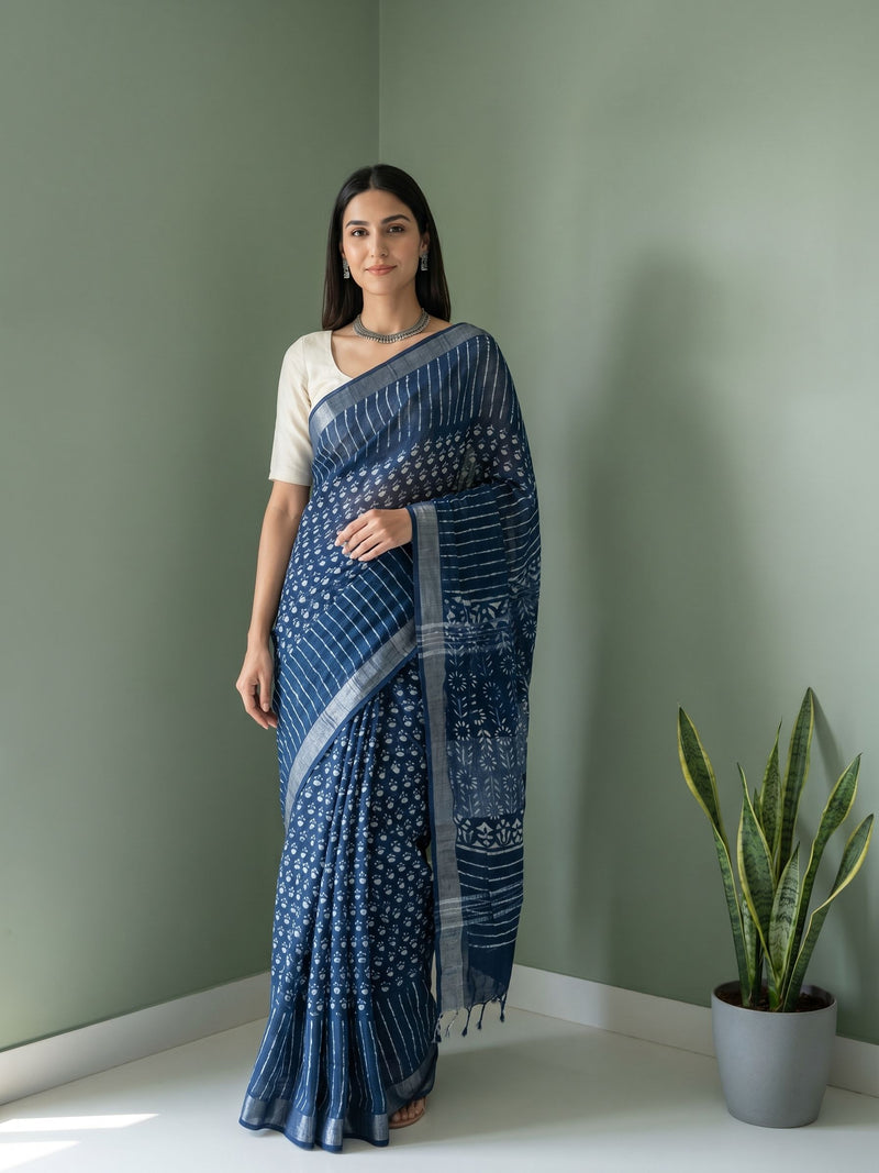 Woman wearing a blue saree standing against a green wall with a plant on the right.