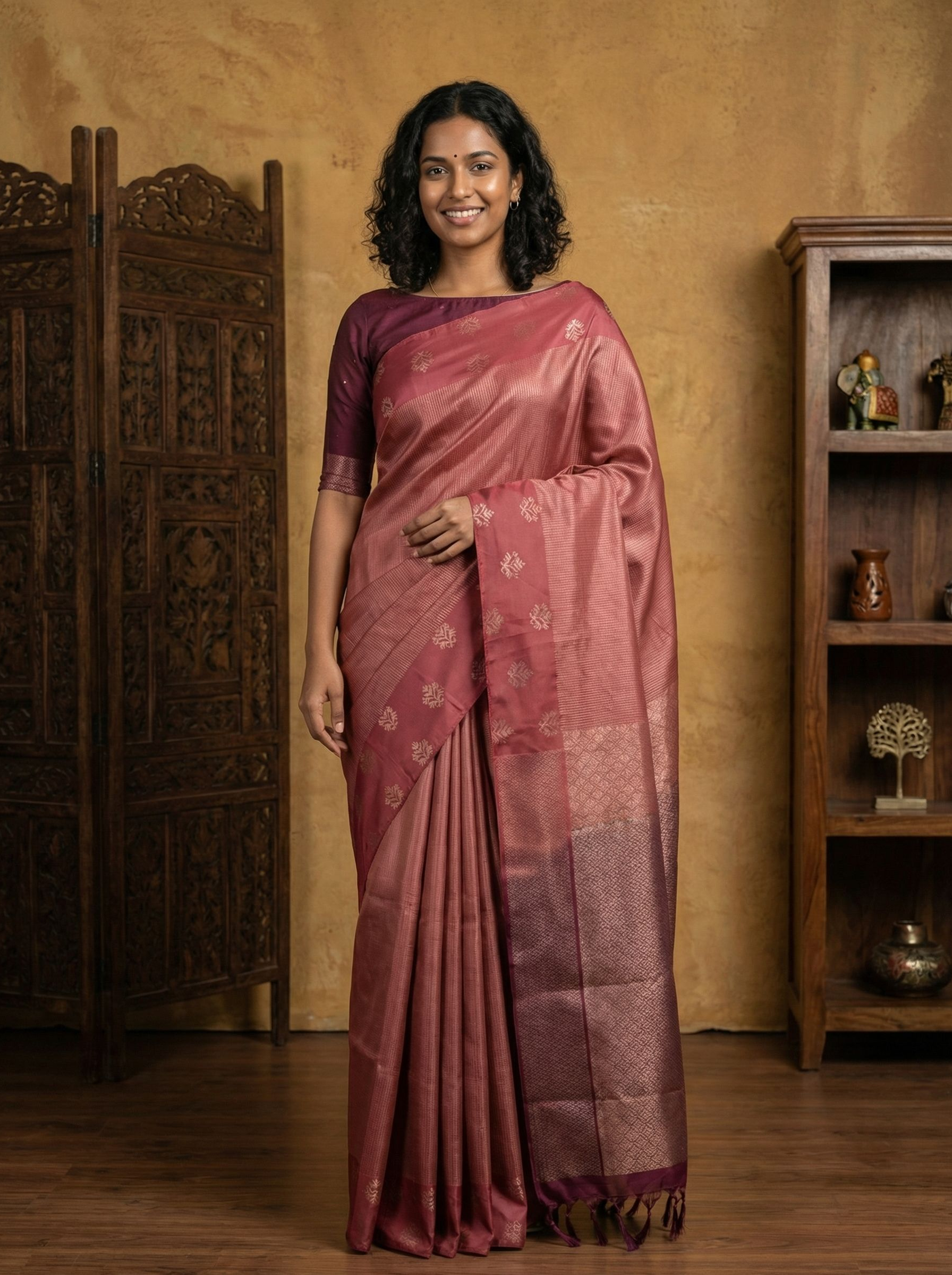Woman wearing a pink saree standing in a room with wooden furniture.