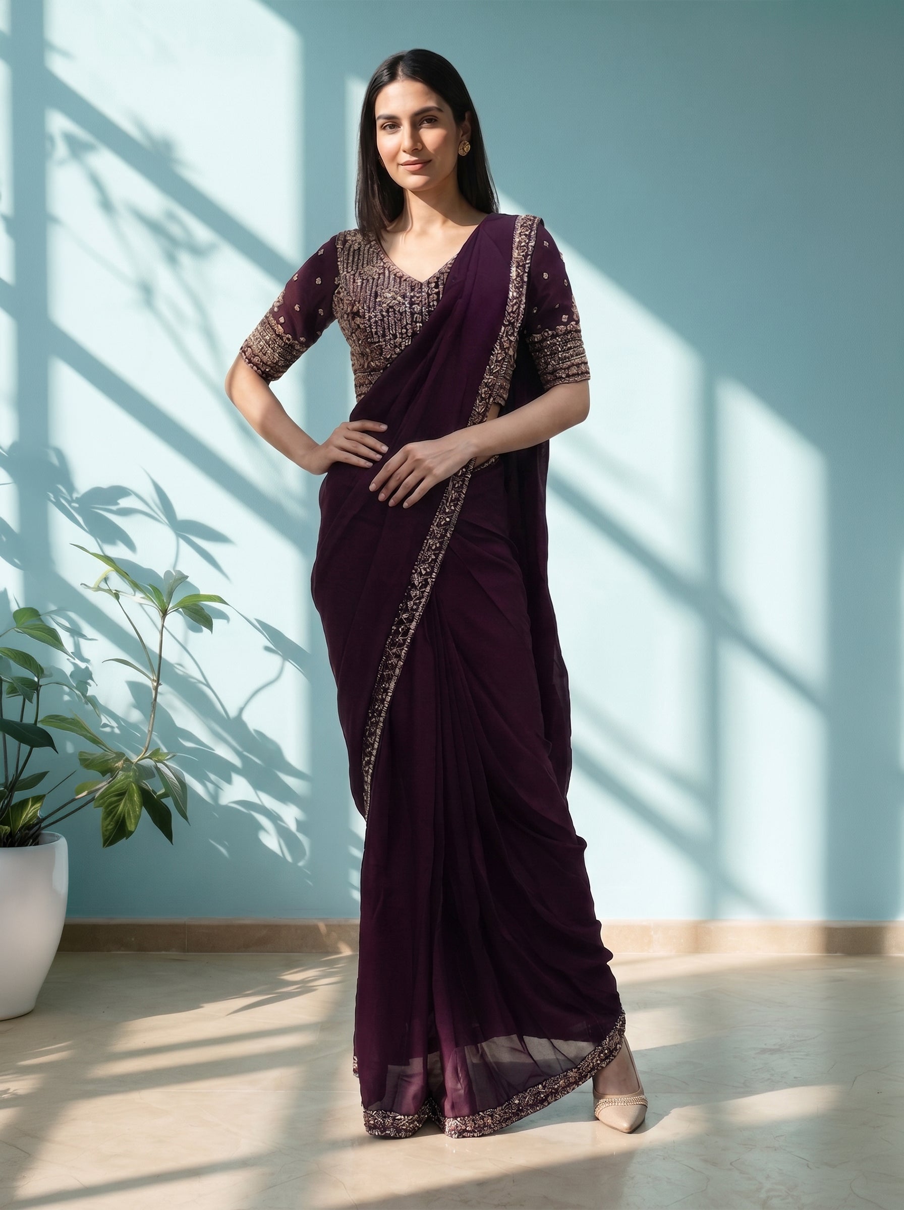 Woman wearing a dark purple saree with gold patterns in a bright room.