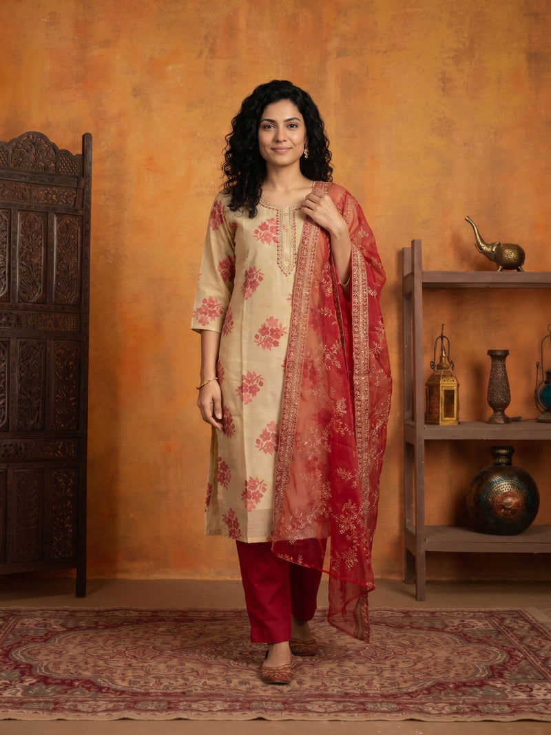 A woman models a traditional Indian salwar kameez suit set consisting of a beige kurta with red floral prints, red pants, and a red dupatta with gold zari embroidery, set against a rustic orange wall with a wooden shelf and carved door.