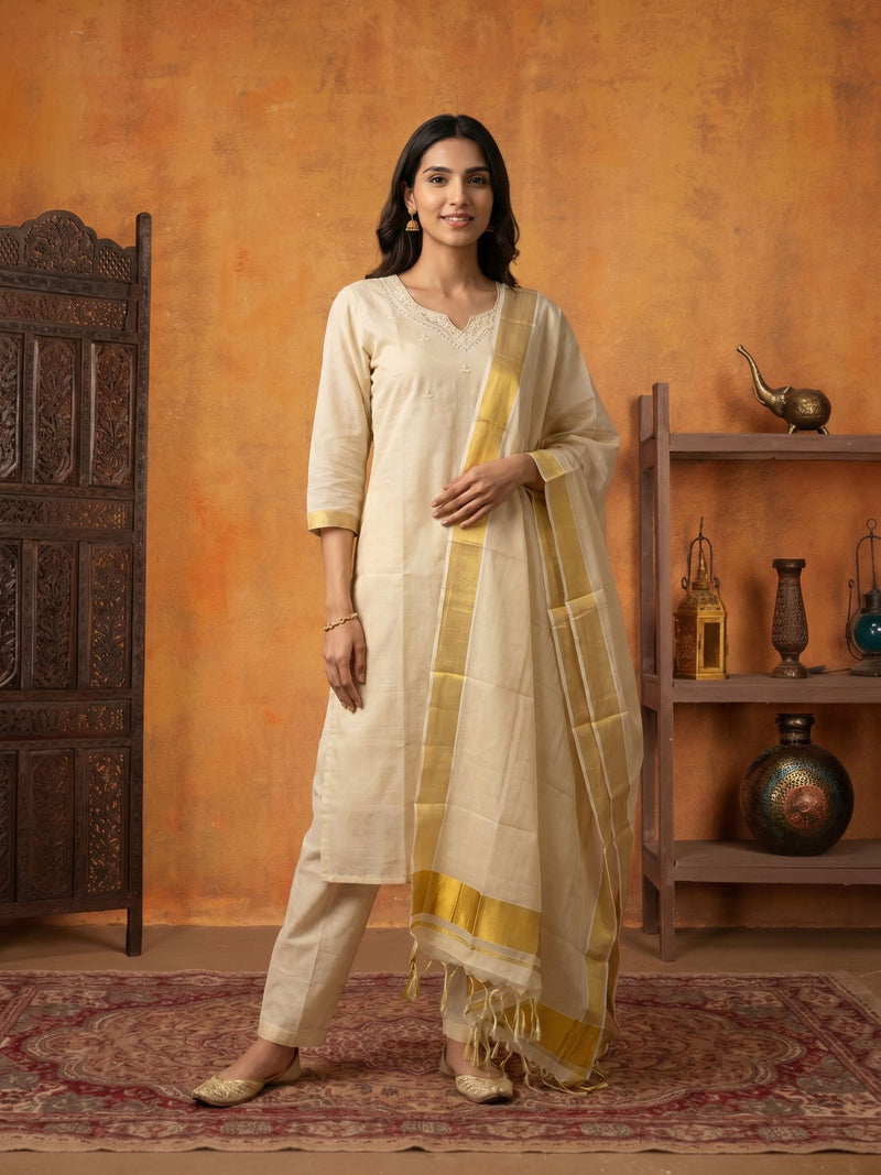 A woman models a traditional Indian outfit consisting of a cream kurta, matching trousers, and a cream dupatta with a yellow border, set against a rustic orange wall with wooden furniture.