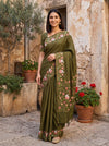 Woman in a green saree with floral patterns standing in front of a stone building with plants.