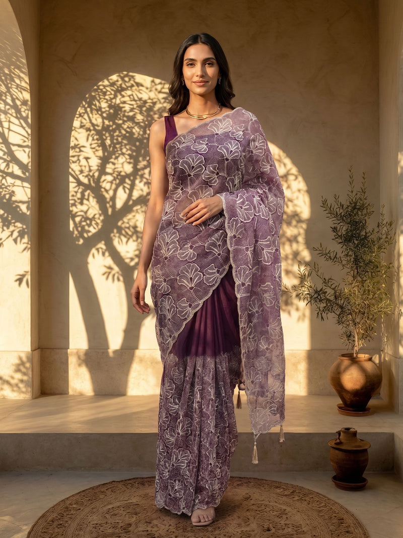 Woman in a purple saree with white patterns standing in a sunlit room.