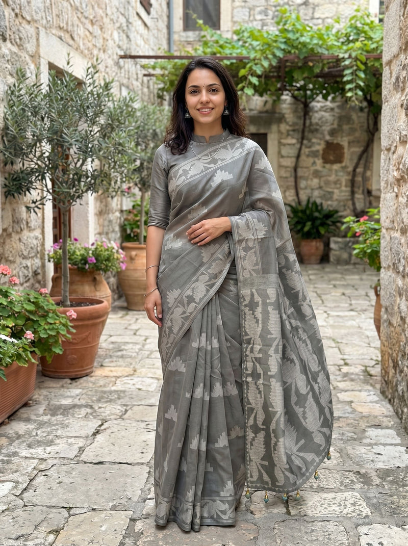 Woman in a gray saree standing in an outdoor setting with stone pavement and plants.
