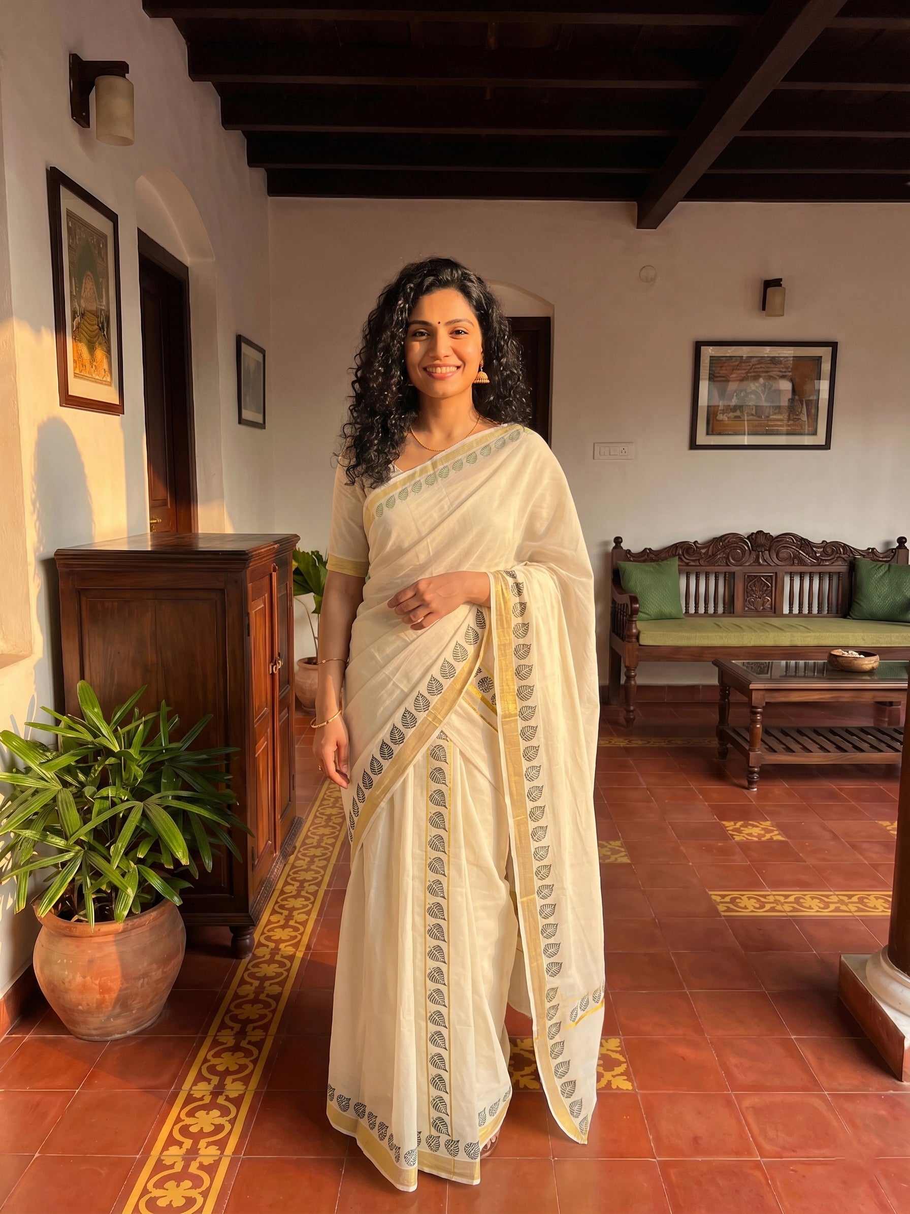 Woman in a white saree standing in a traditional room with wooden furniture and decor.