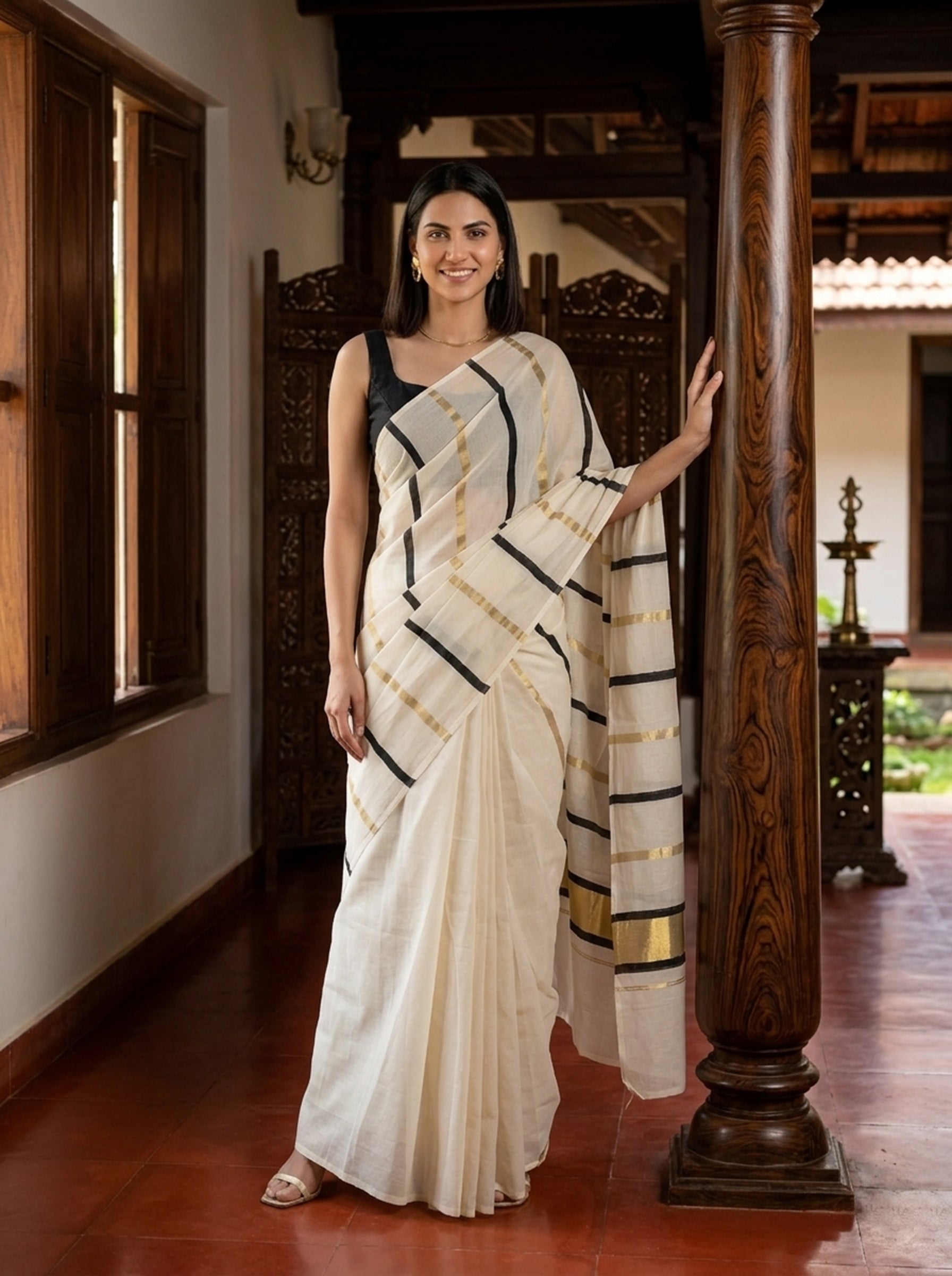 Woman in a white saree with black and gold patterns standing in a traditional setting.