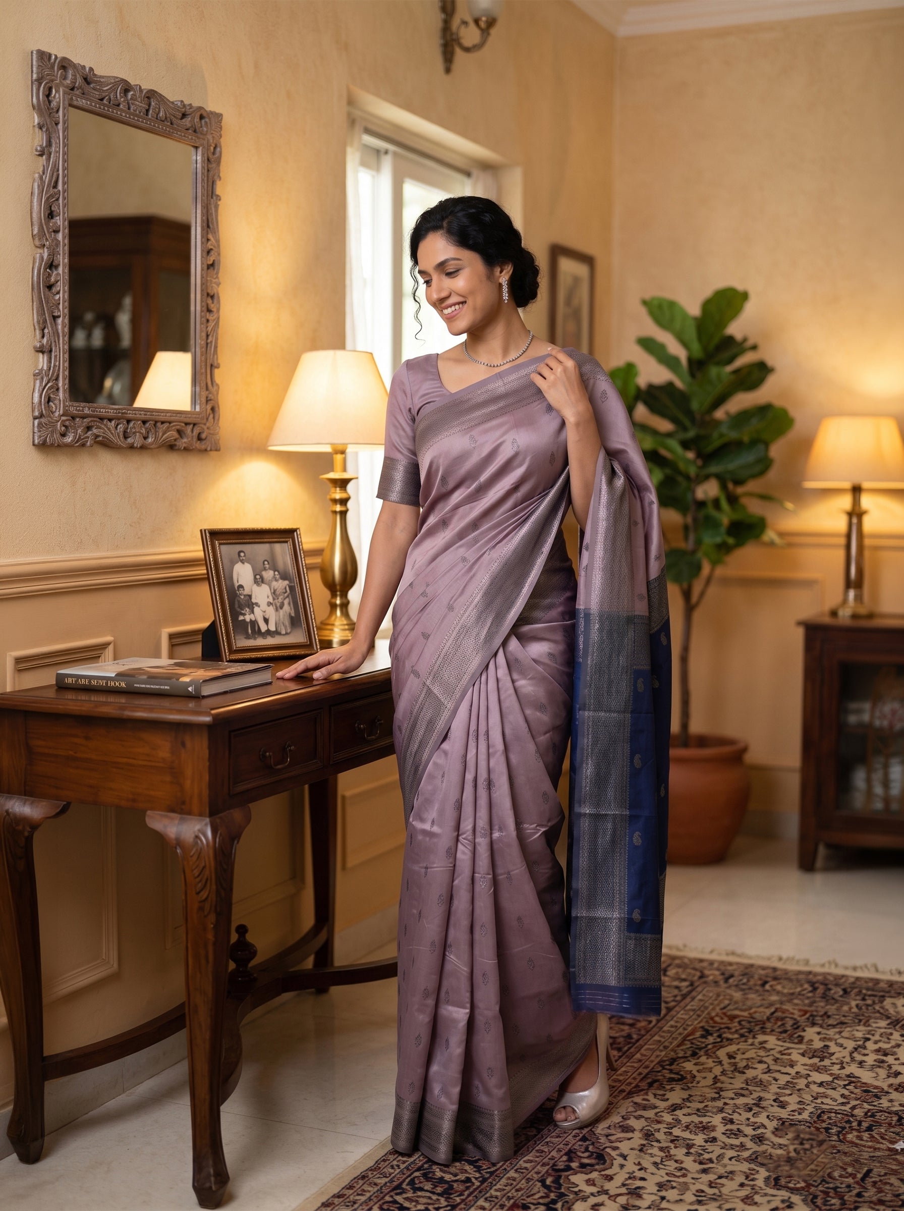 Woman in a lavender saree standing in a room with a desk and lamp.