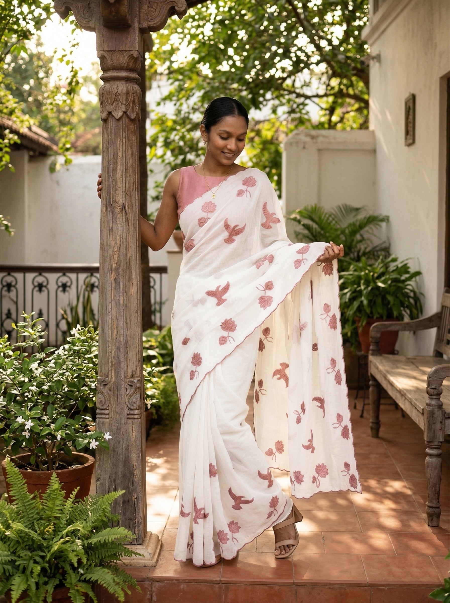 Woman in a white saree with pink floral patterns standing outdoors near a wooden pillar.