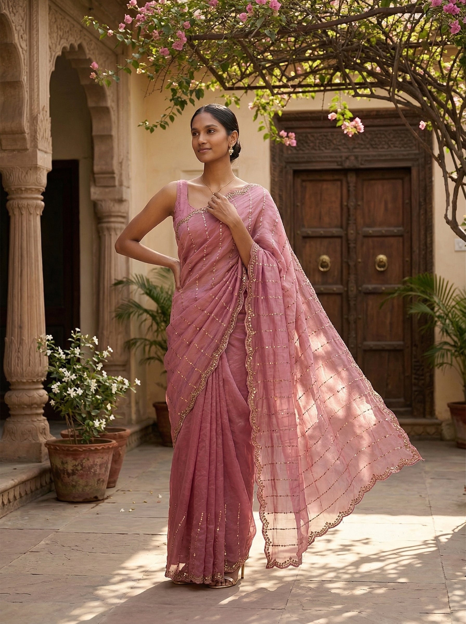 Woman in a pink saree standing in a courtyard with floral decorations.