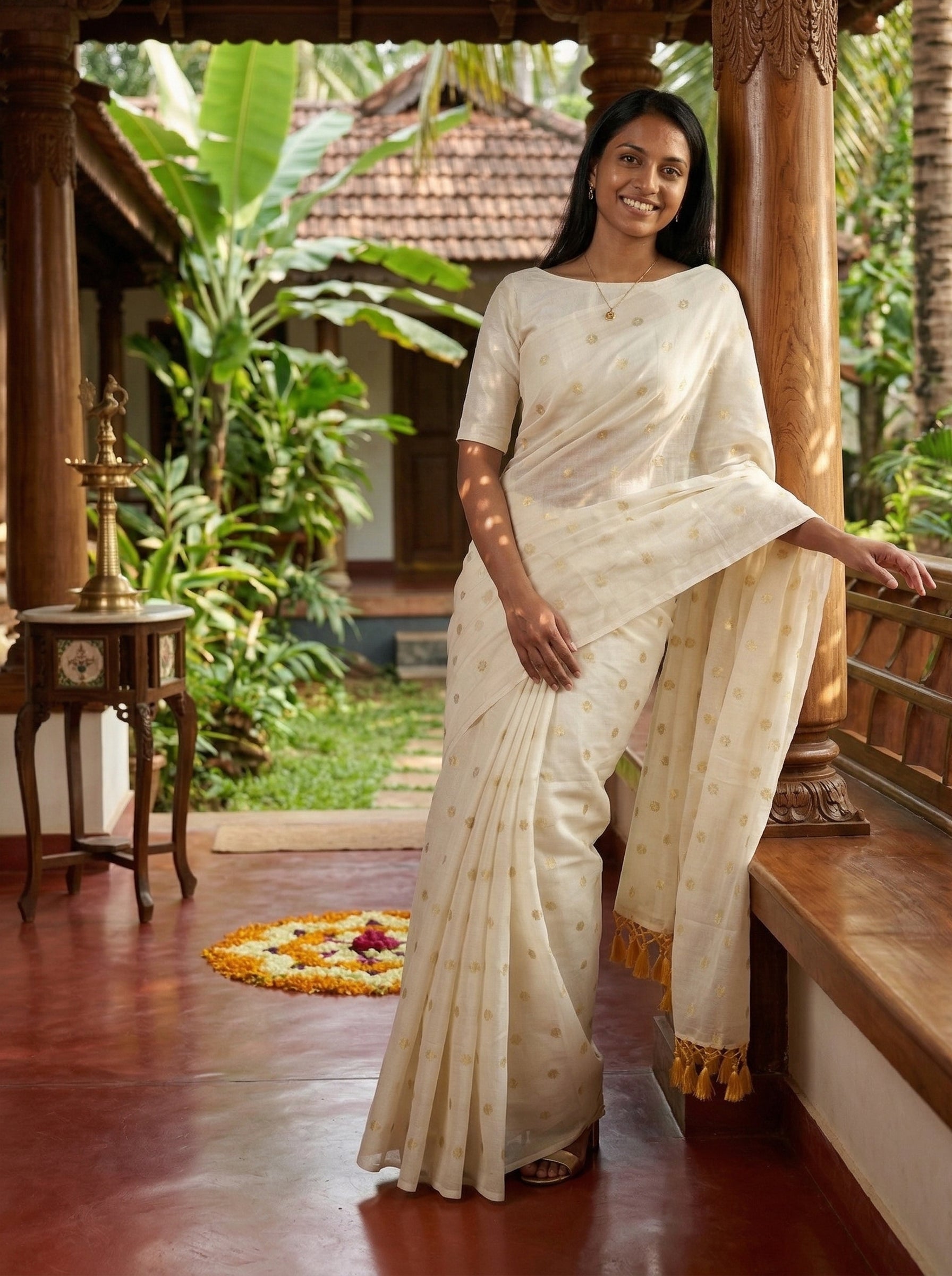 Woman in a white saree standing in a traditional setting with greenery.