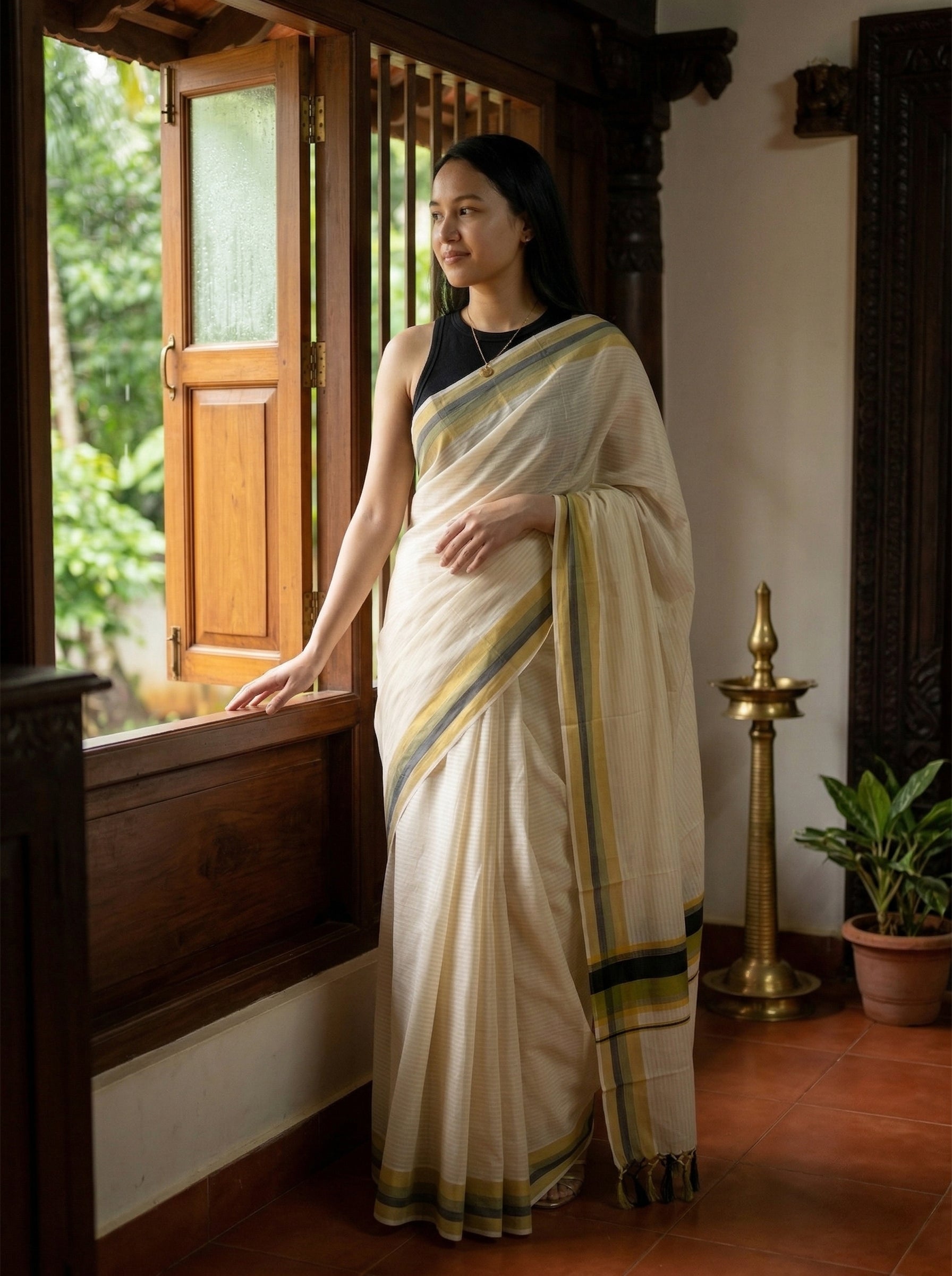 Woman in a white saree with green and yellow stripes standing by a window.