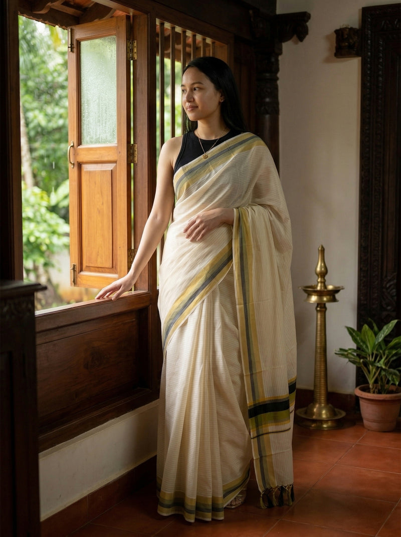 Woman in a white saree with green and yellow stripes standing by a window.