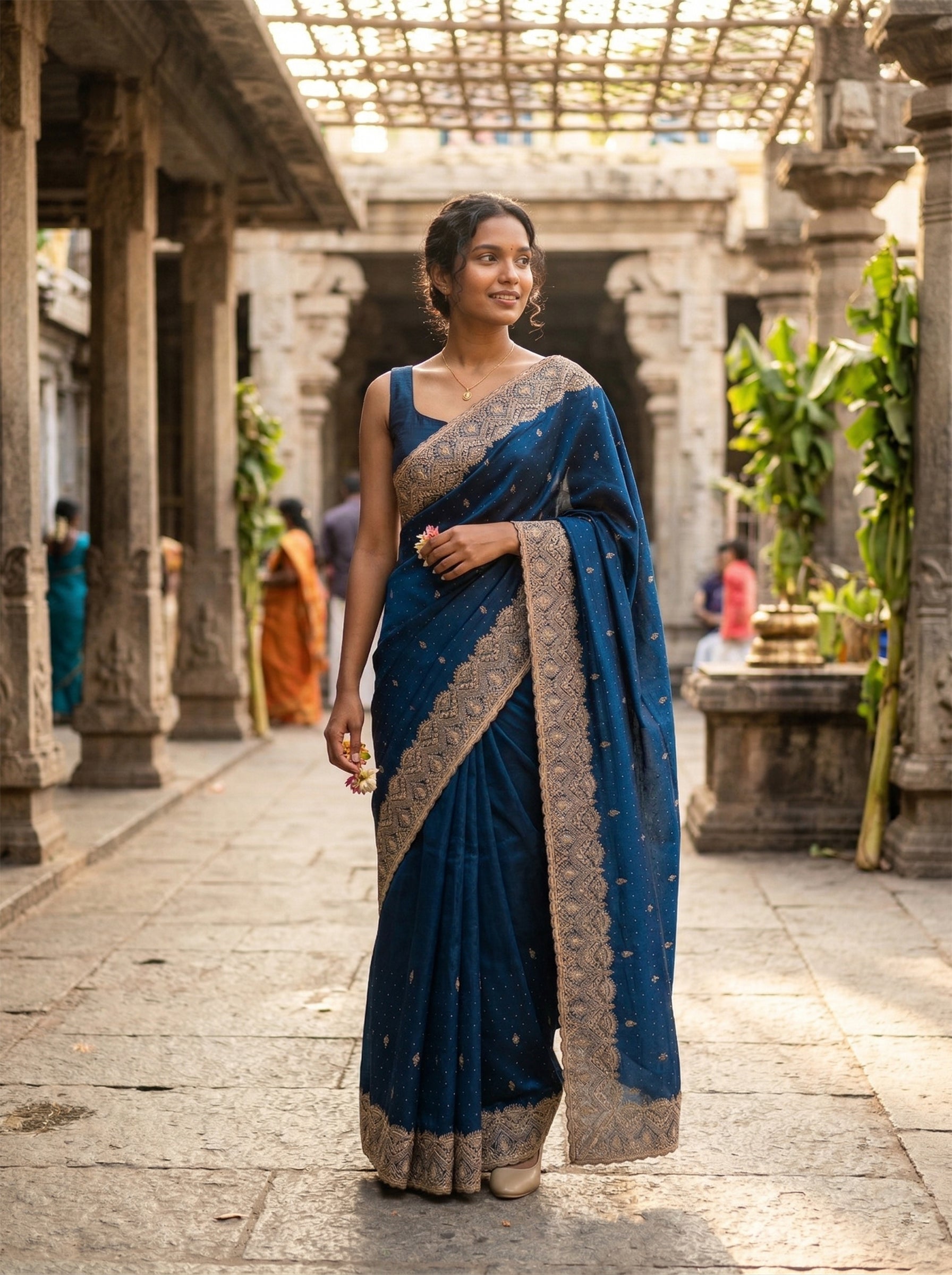 Woman in a blue saree standing in an outdoor setting with stone architecture and plants.