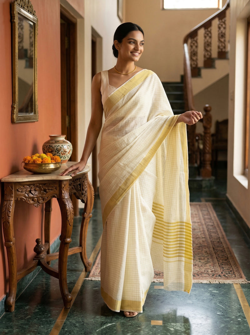 Woman in a white saree with yellow border standing in a decorated room.