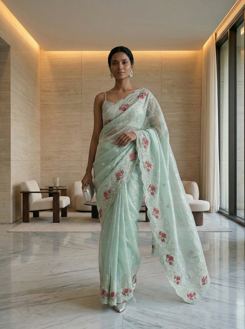 Woman in a light green saree with floral patterns standing in an elegant indoor setting.