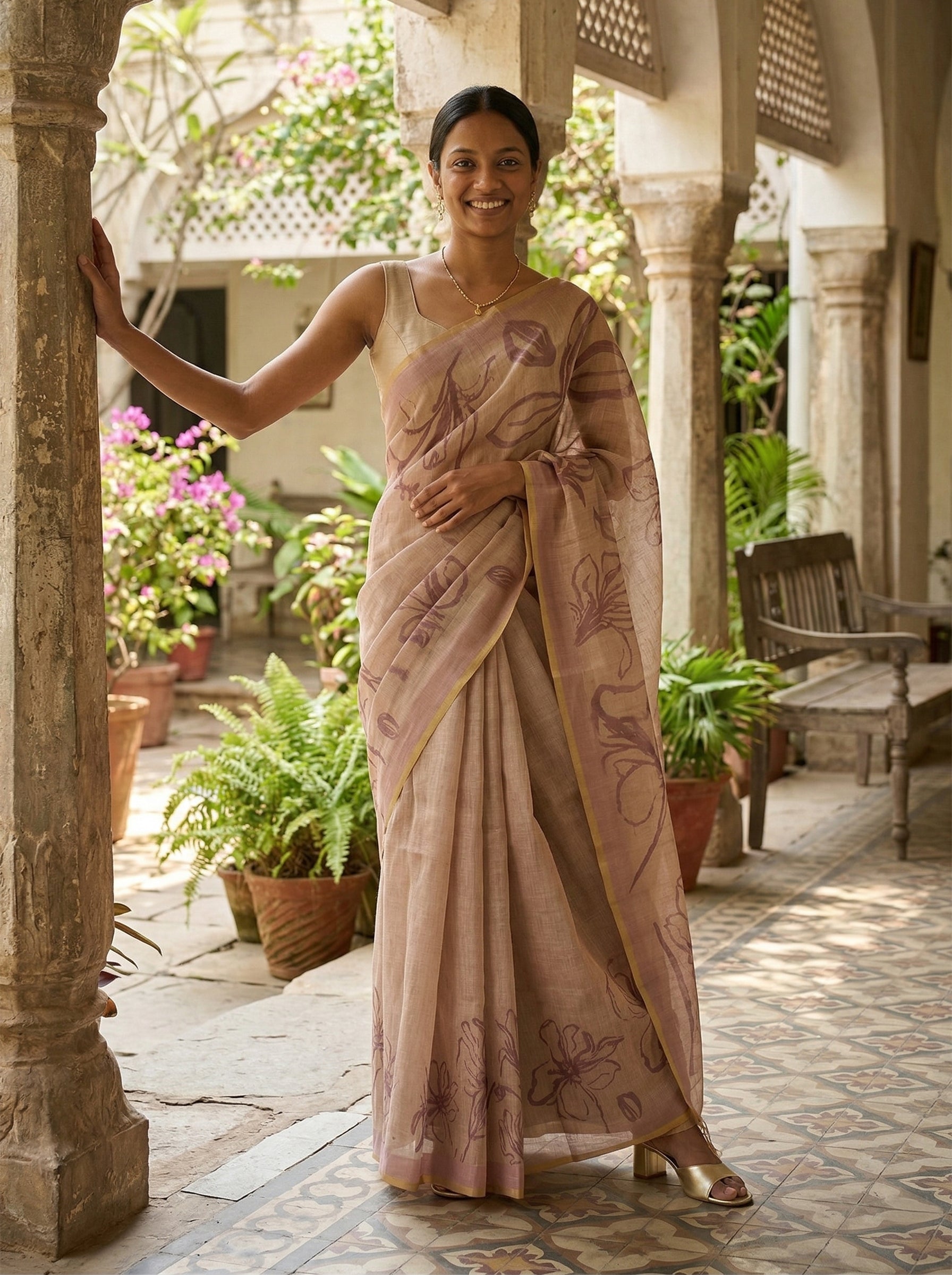 Woman in a traditional saree standing in an outdoor setting with plants and architectural elements.