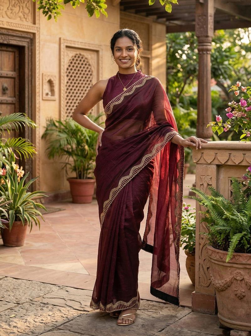 Woman in a maroon saree standing in an outdoor setting with plants and a building.