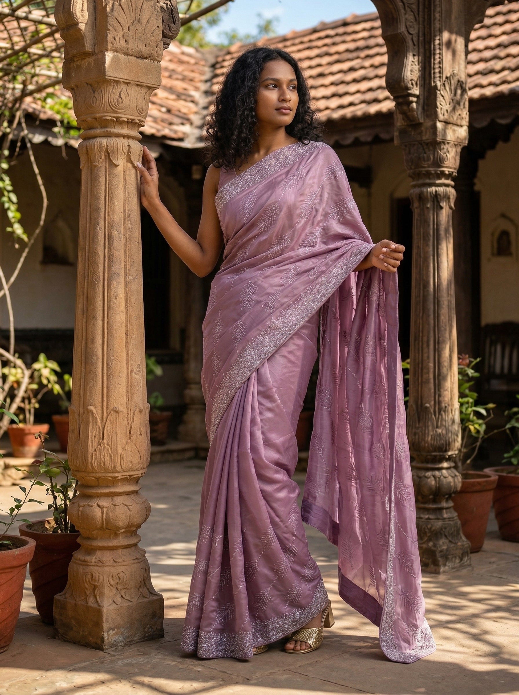 Woman in a pink saree standing in an outdoor setting with architectural columns and plants.