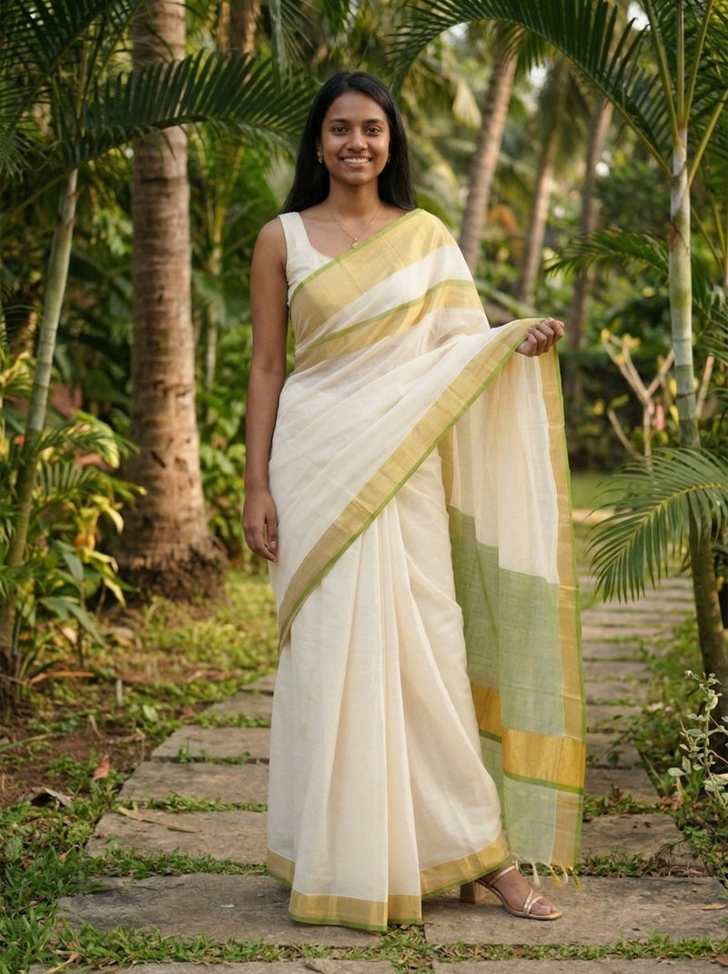 Woman in a white saree with green and yellow border standing in a tropical setting.