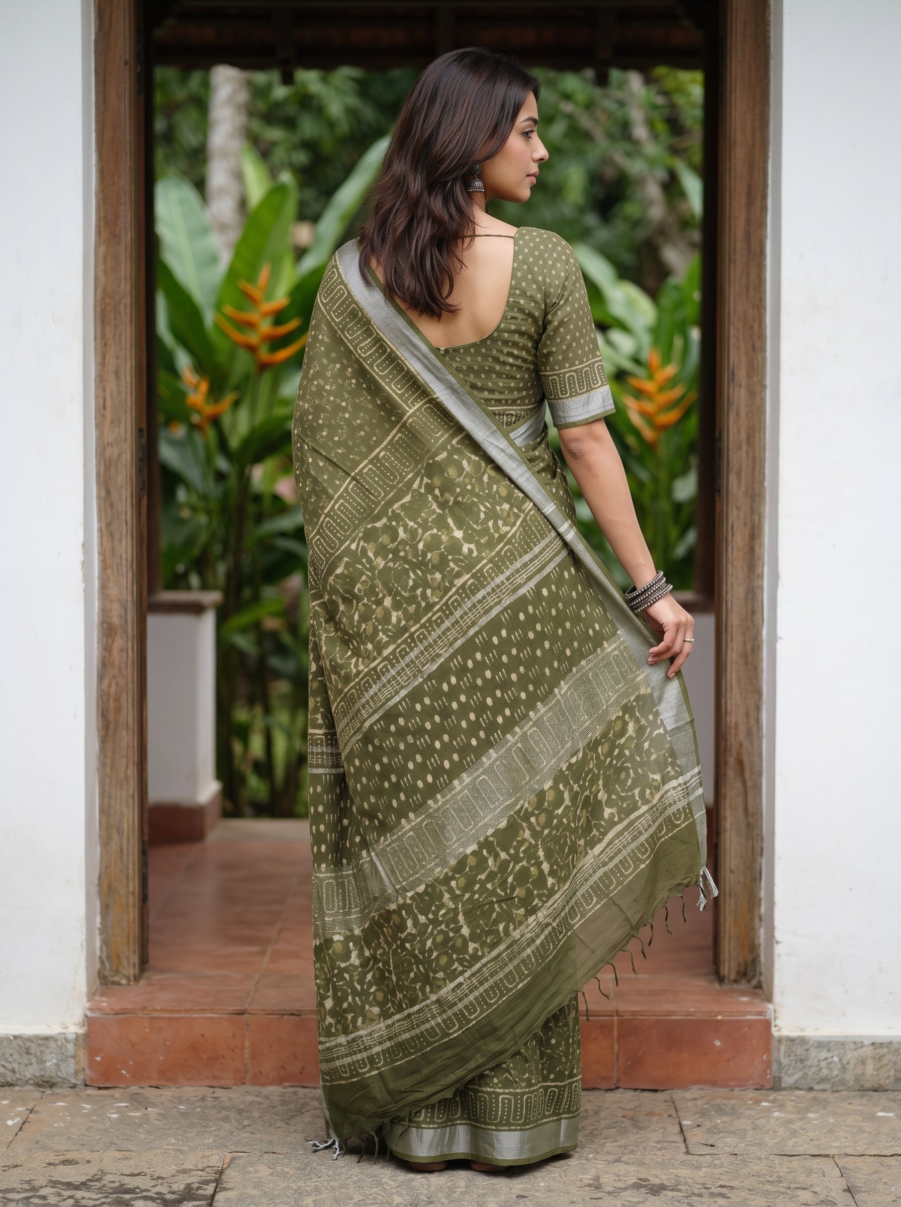A woman wearing an olive green printed saree with a matching printed blouse, standing in a doorway with lush green plants visible in the background.