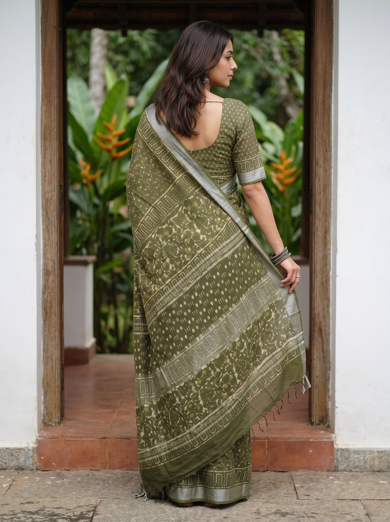A woman wearing an olive green printed saree with a matching printed blouse, standing in a doorway with lush green plants visible in the background.