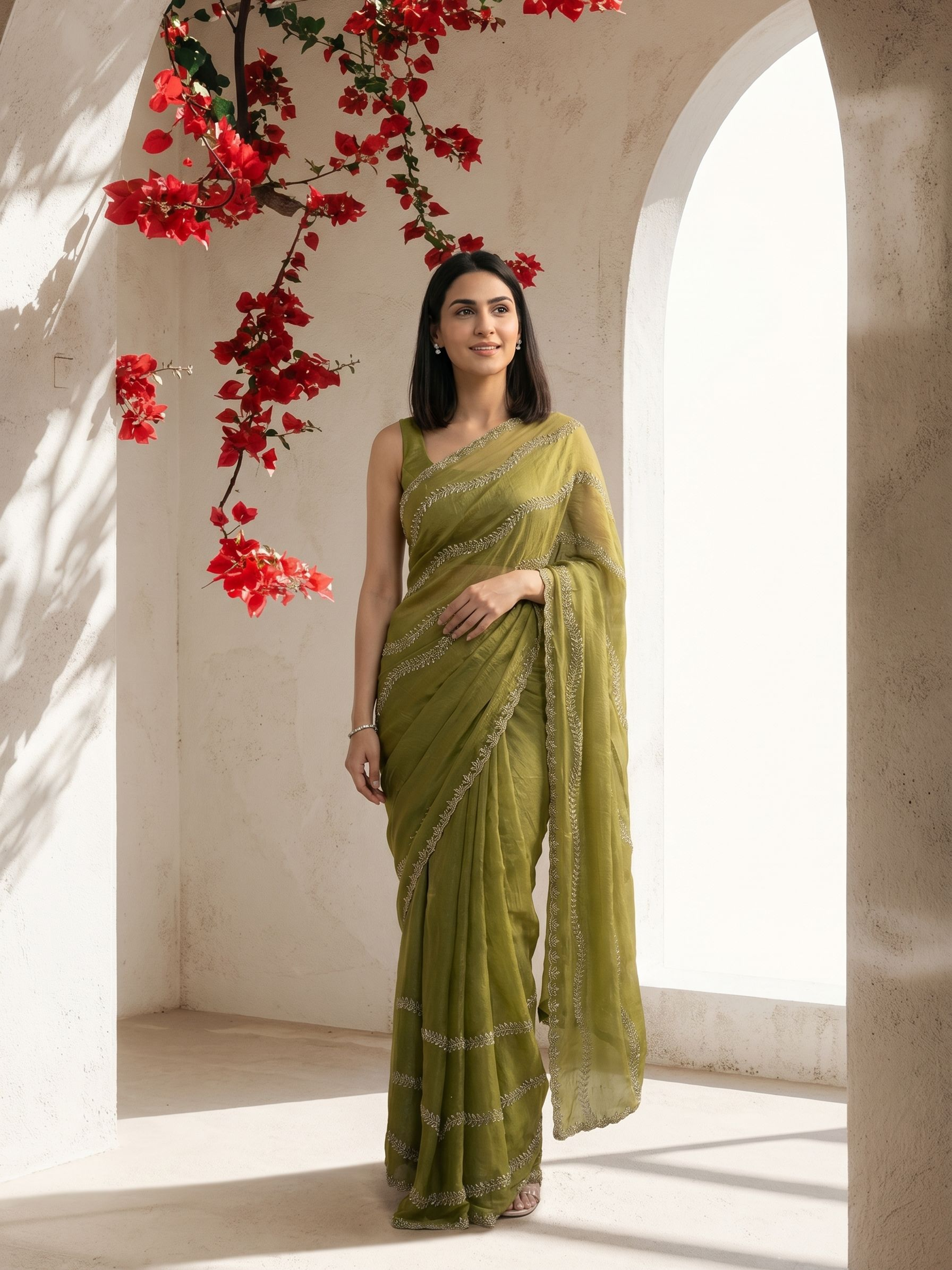 Woman in a green saree standing in a sunlit room with red flowers.